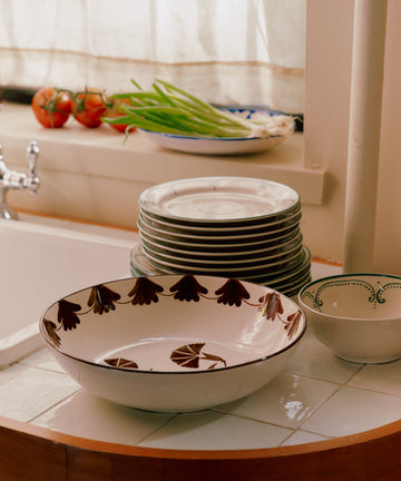 The Catalina Serving Bowl sits on the tiled kitchen counter beside a stack of plates, with tomatoes and green onions arranged nearby and sunlight streaming in through the window.