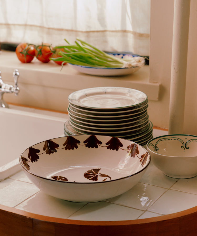 The Catalina Serving Bowl sits on the tiled kitchen counter beside a stack of plates, with tomatoes and green onions arranged nearby and sunlight streaming in through the window.