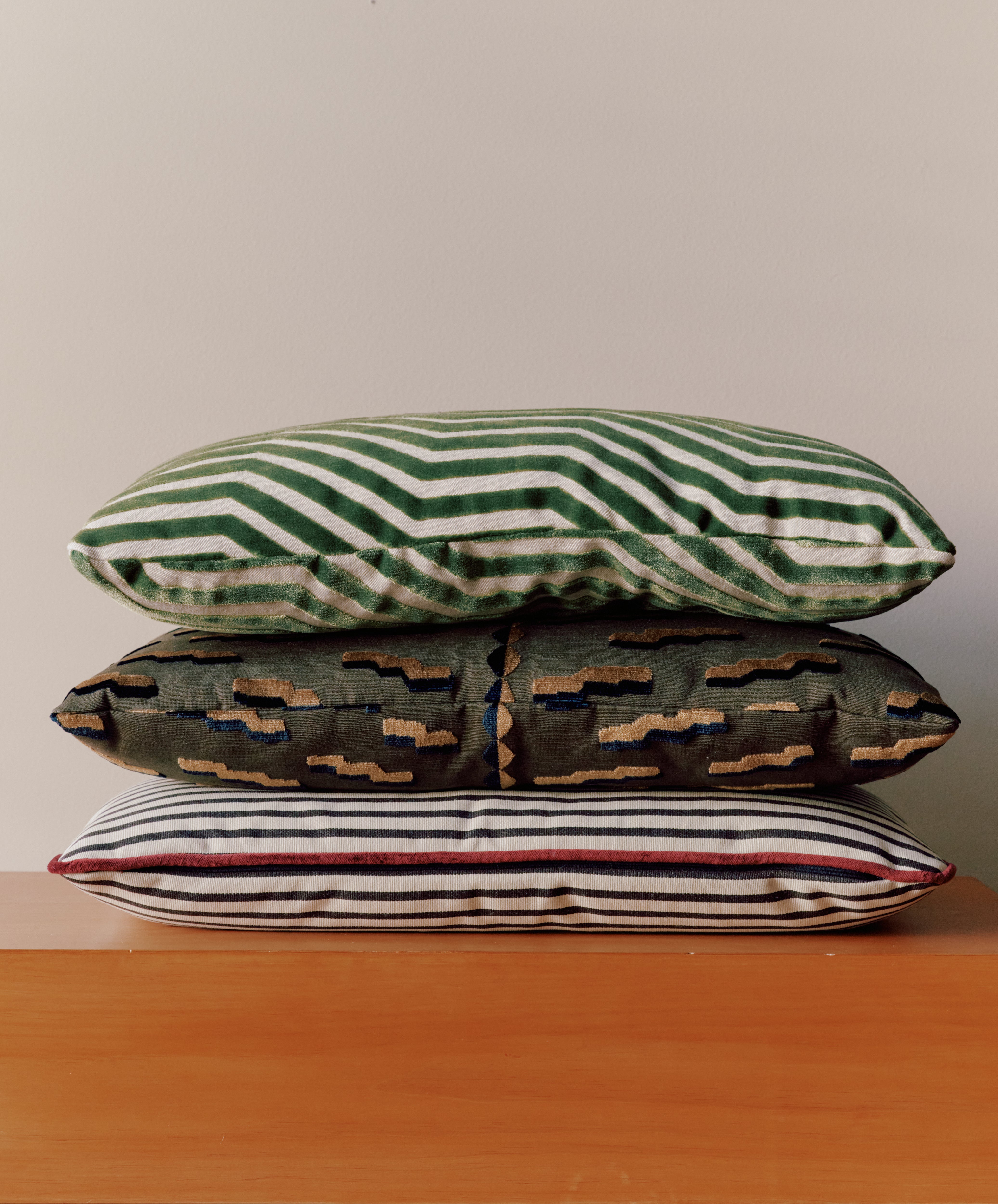 The Polo Lumbar Cushion, crafted from dead-stock fabric, features bold black stripes and is displayed stacked with two other patterned pillows on a wooden surface against a plain light wall.