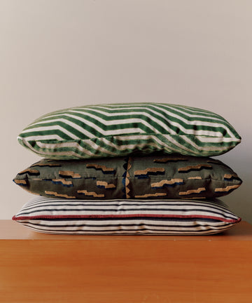 The Polo Lumbar Cushion, crafted from dead-stock fabric, features bold black stripes and is displayed stacked with two other patterned pillows on a wooden surface against a plain light wall.