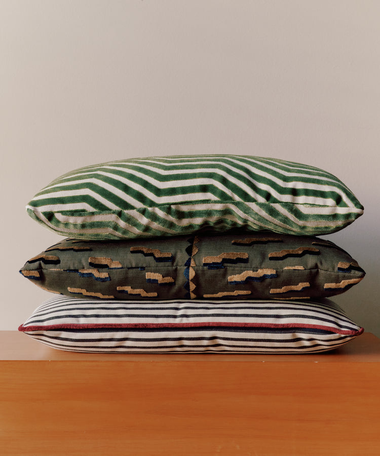 The Polo Lumbar Cushion, crafted from dead-stock fabric, features bold black stripes and is displayed stacked with two other patterned pillows on a wooden surface against a plain light wall.