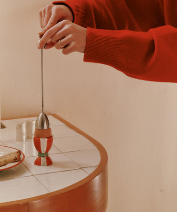 A person in a red sweater uses the Clack Egg Opener on a soft-boiled egg in an egg cup, placed on a tiled countertop beside a plate of bread.