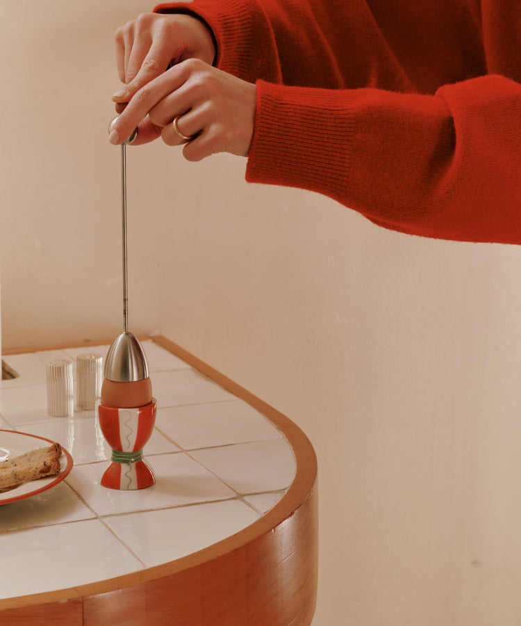 A person in a red sweater uses the Clack Egg Opener on a soft-boiled egg in an egg cup, placed on a tiled countertop beside a plate of bread.