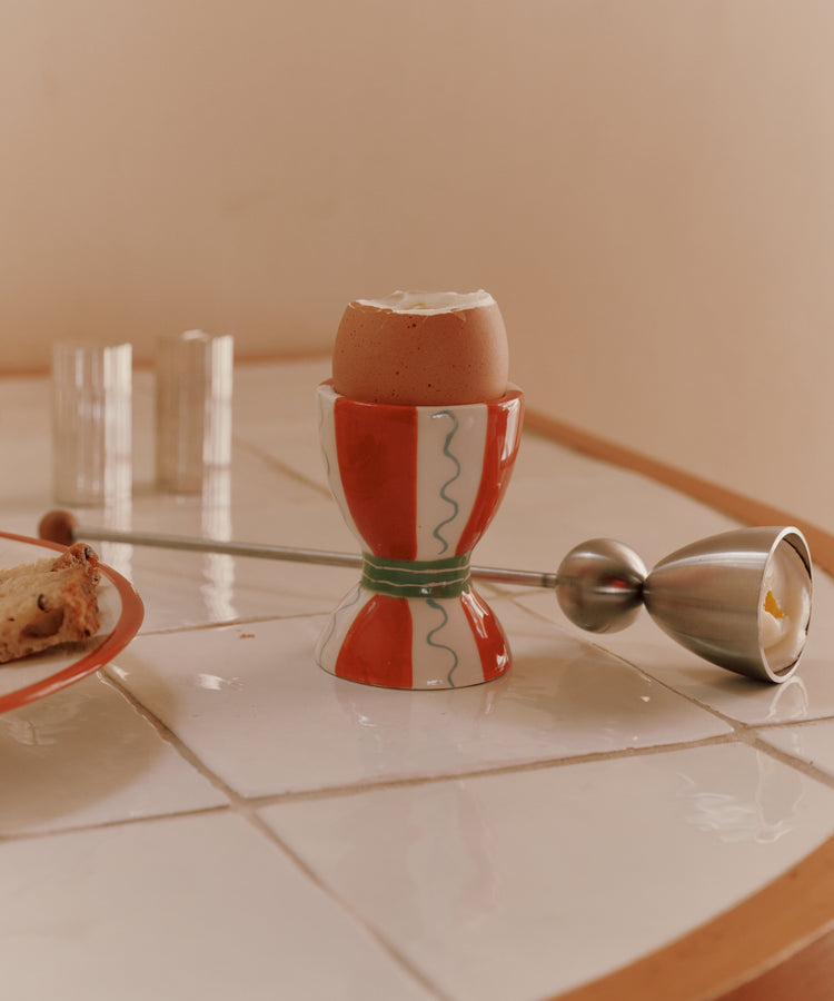 A soft-boiled egg in a red and white egg cup is on a tiled table beside the German-engineered Clack Egg Opener and a plate with a piece of bread.