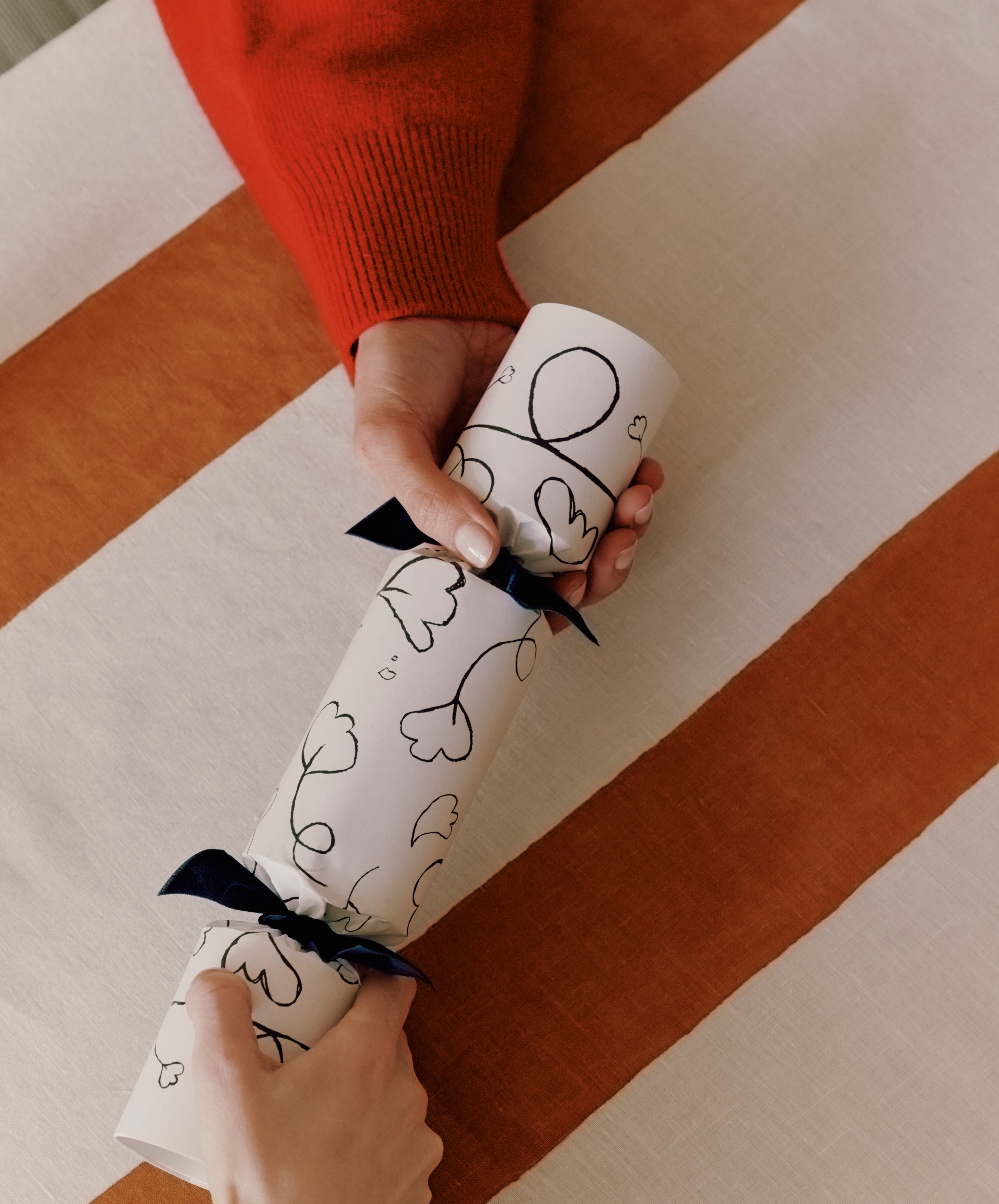 Two people hold opposite ends of a Capriccio Christmas Cracker with a ribbon, positioned over a striped tablecloth.