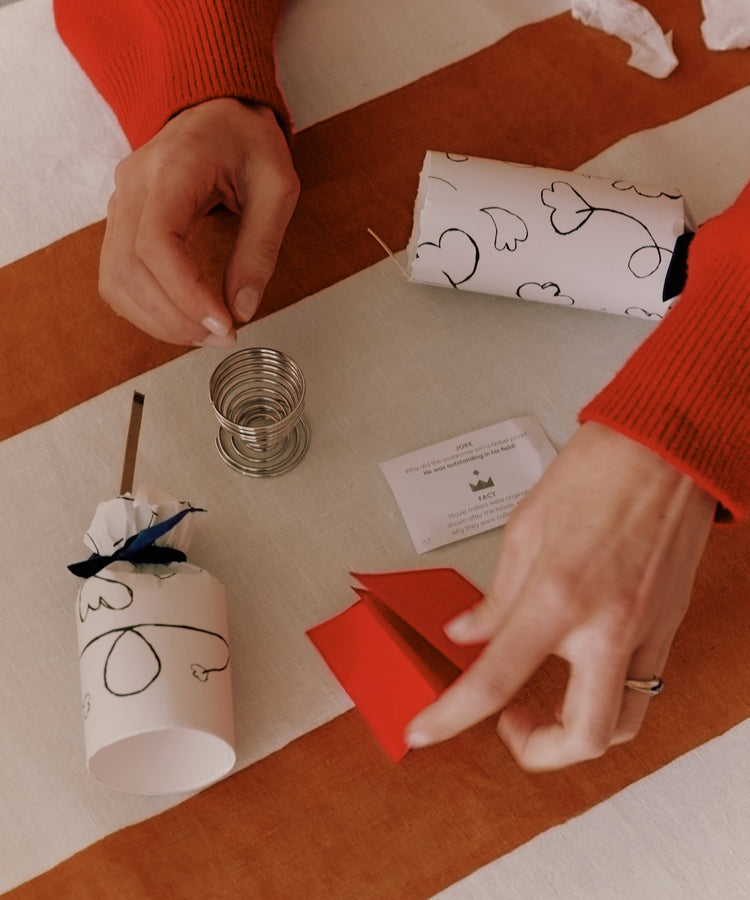 Two hands open a white Capriccio Christmas Cracker with black heart designs on a striped tablecloth; inside are a metal spring, small card, and red paper fortune—ideal festive party favors or Christmas crackers.
