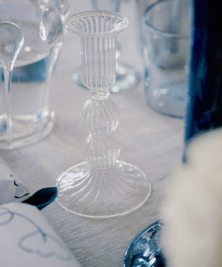 The Elsa Candleholder, a clear hand-blown piece with a ribbed design, sits on a white tablecloth among glassware, bringing timeless elegance to the setting.