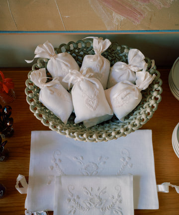 A woven basket holds six Lavender Sachets, handmade in Portugal with Viana do Castelo embroidery, each tied with ribbons and displayed on a wooden surface beside embroidered linens.
