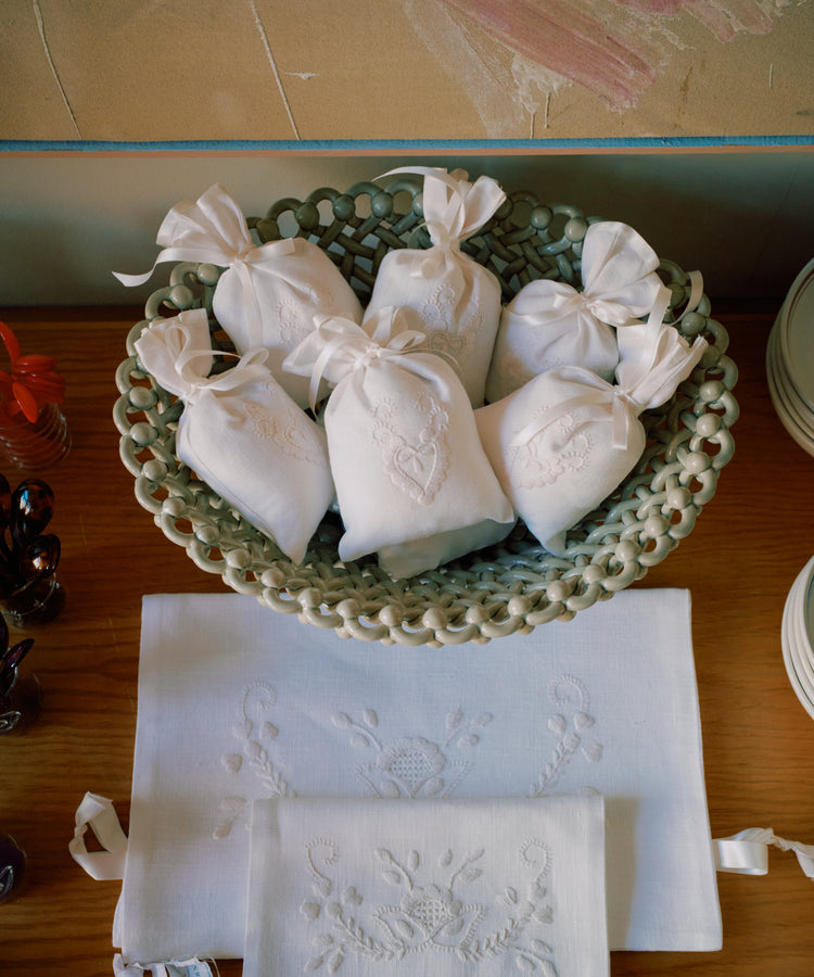 A woven basket holds six Lavender Sachets, handmade in Portugal with Viana do Castelo embroidery, each tied with ribbons and displayed on a wooden surface beside embroidered linens.