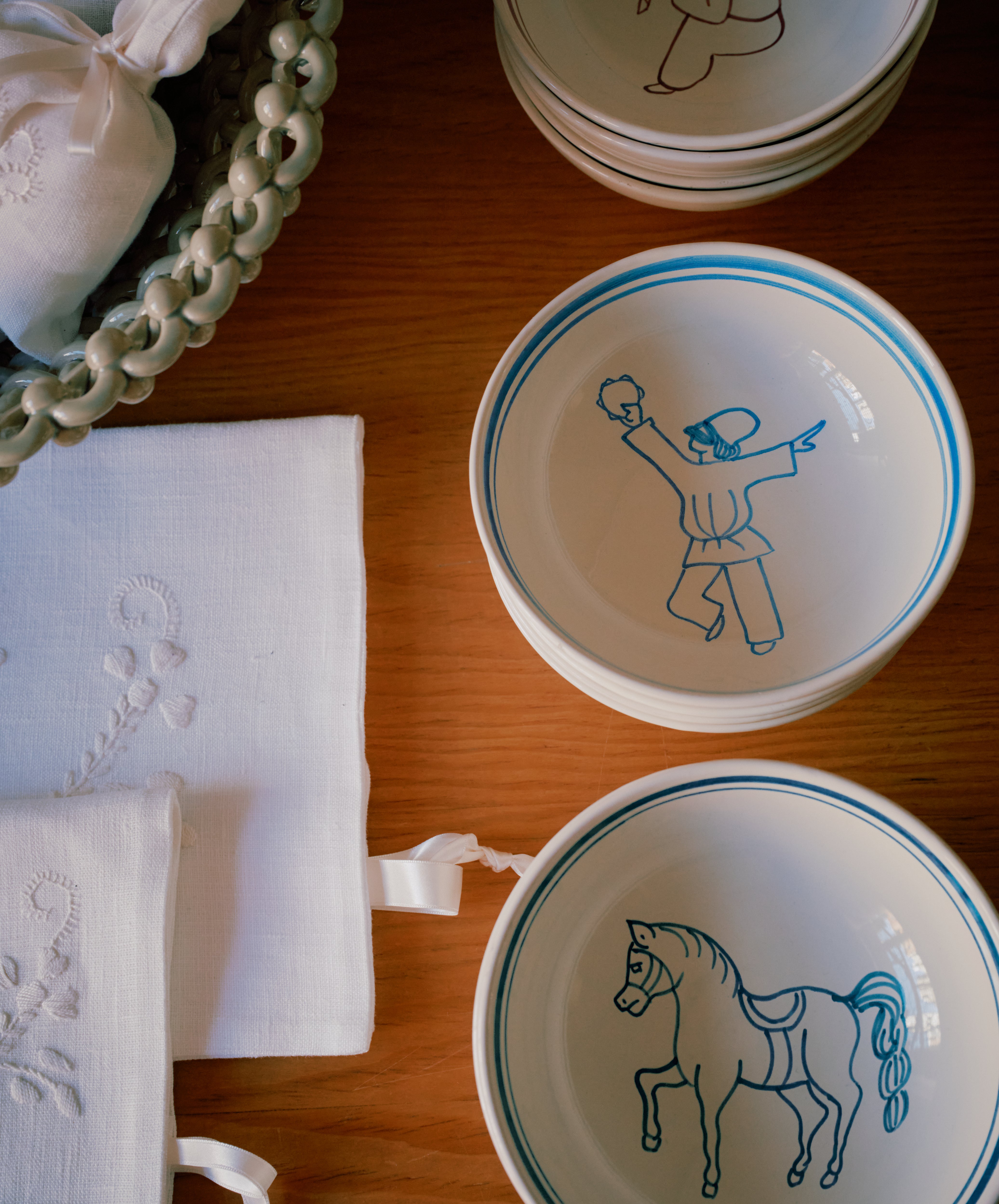 The Folk Cereal Bowl, Jester features whimsical blue line art of a person and a horse, hand-painted by an Italian ceramist; it’s displayed on a wooden table beside a basket and white embroidered cloths.