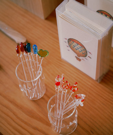 Two glass cups hold Heart Cocktail and Nibble Sticks—one set with colorful shapes, the other with red-tipped hearts—beside a stack of hot air balloon cards, making for charming drink accessories.