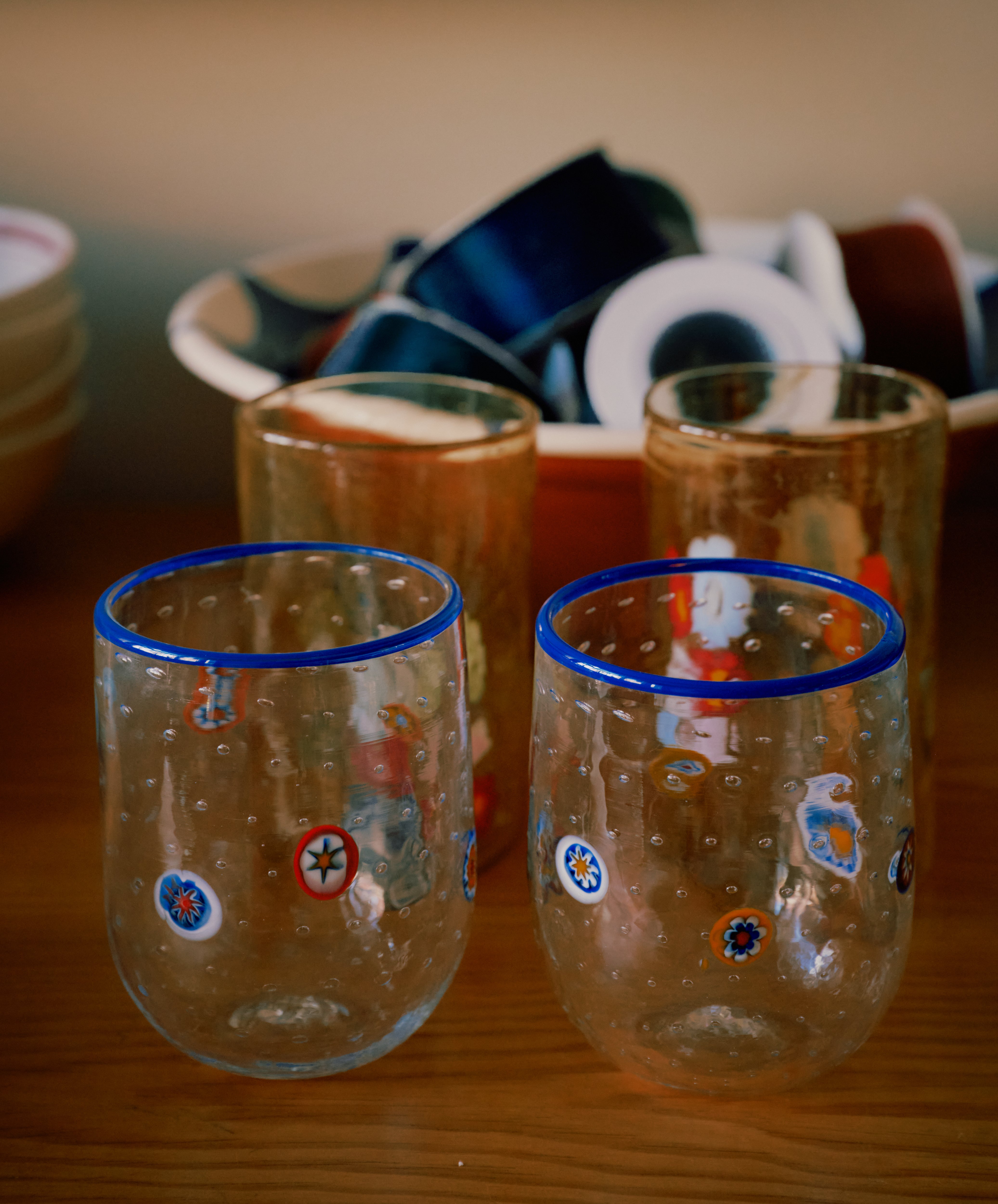 A set of two Girotondo Glasses with blue rims and colorful patterns showcases Venetian craftsmanship on a wooden surface, with bowls and small dishes in the background.
