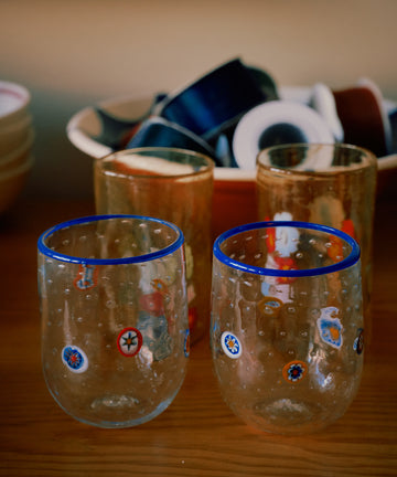 A set of two Girotondo Glasses with blue rims and colorful patterns showcases Venetian craftsmanship on a wooden surface, with bowls and small dishes in the background.
