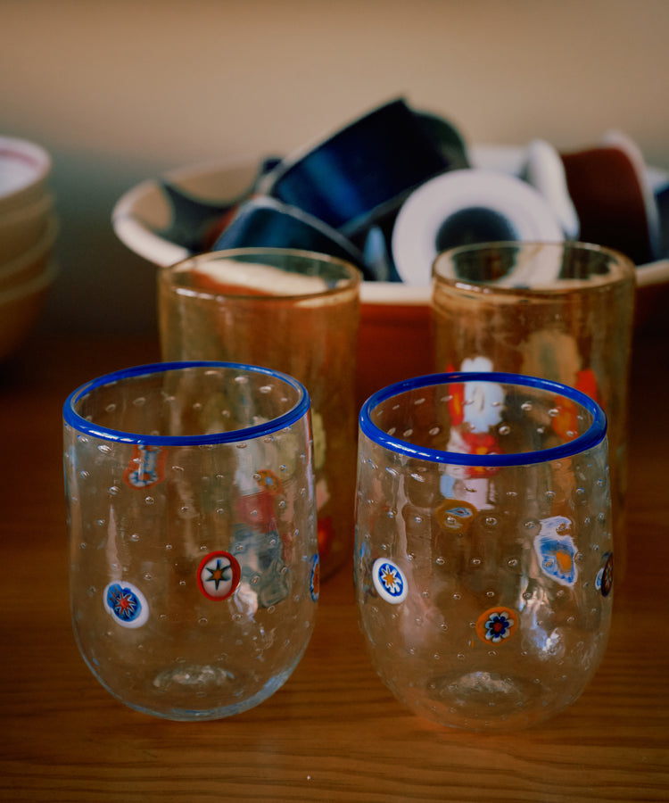 A set of two Girotondo Glasses with blue rims and colorful patterns showcases Venetian craftsmanship on a wooden surface, with bowls and small dishes in the background.