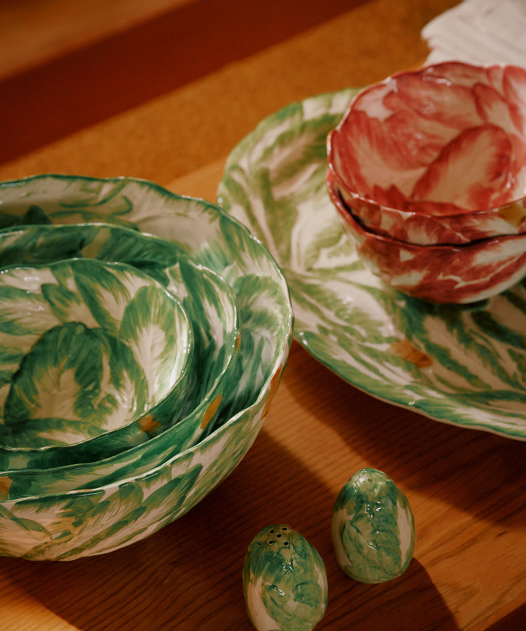 The Radicchio Serving Bowls, adorned with hand-painted green and red leaf patterns, are nested on a wooden table alongside matching salt and pepper shakers—ideal Italian tableware for any gathering.