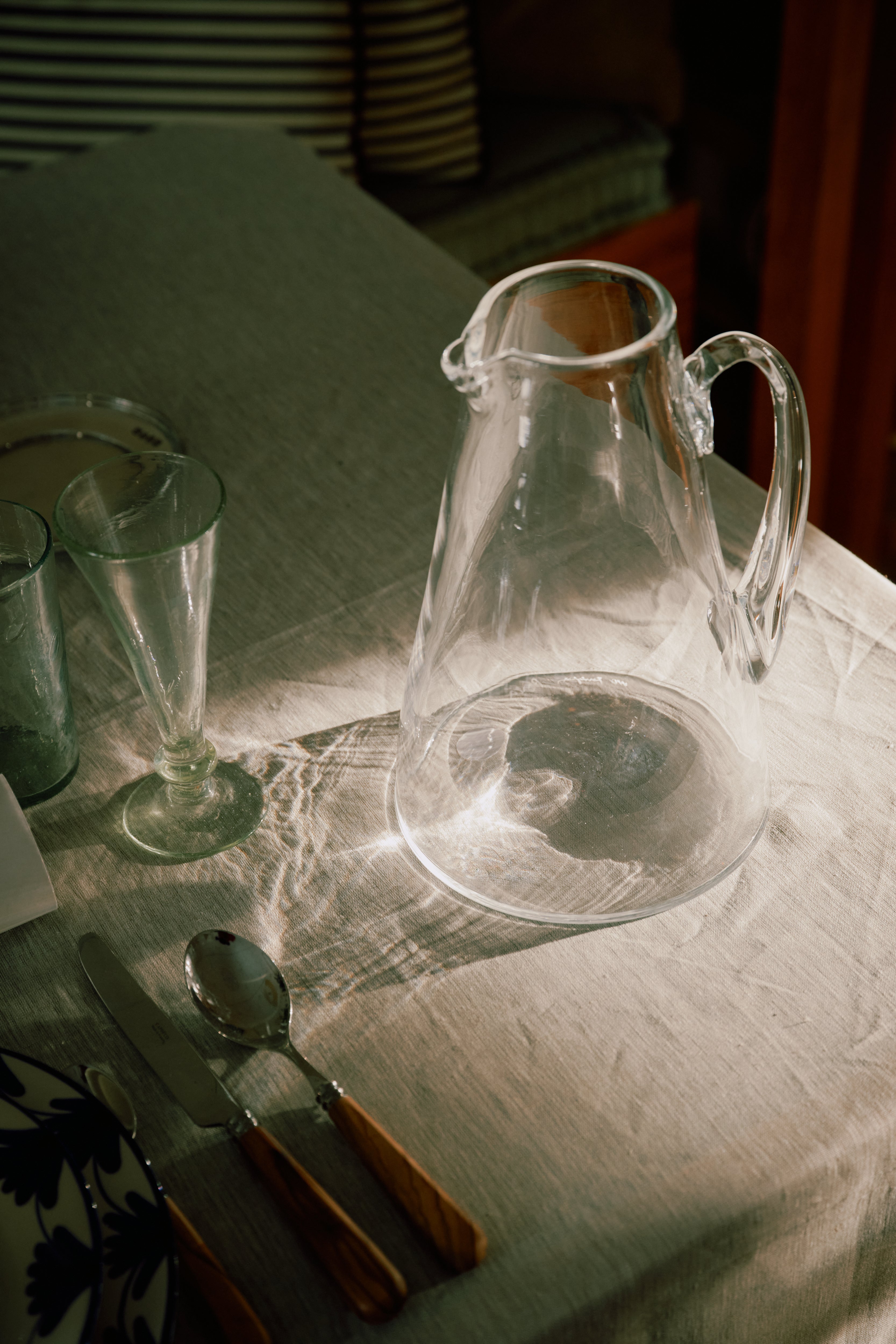The Gemma Jug, made of clear glass with crystal transparency, sits on a sunlit tablecloth beside a spoon, knife, fork, empty glass, and patterned plate.
