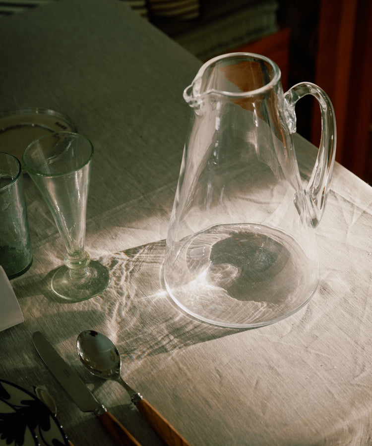 The Gemma Jug, made of clear glass with crystal transparency, sits on a sunlit tablecloth beside a spoon, knife, fork, empty glass, and patterned plate.