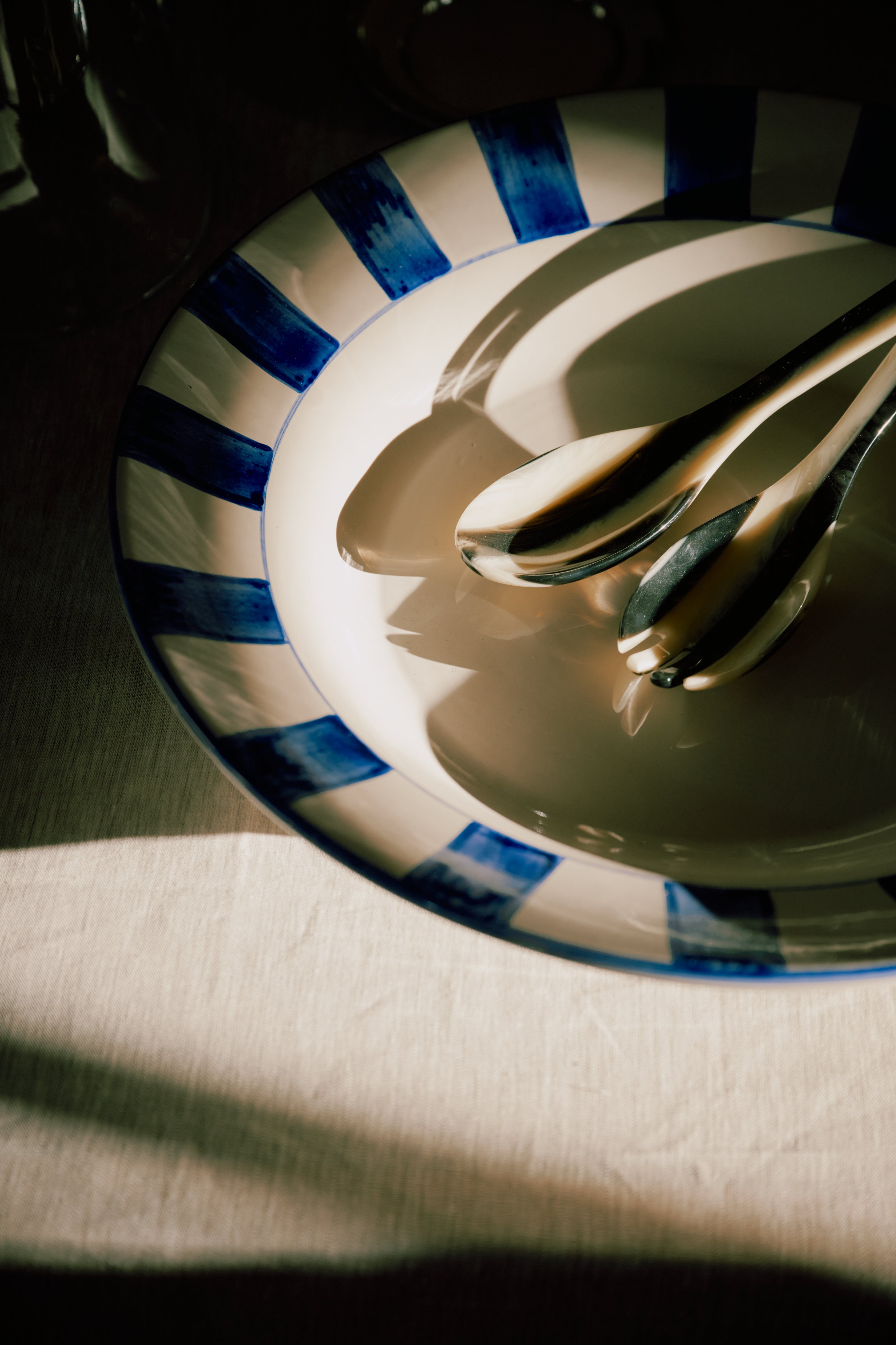 The Bestia Serving Set, featuring two metal spoons, rests on a ceramic plate with blue stripes and casts shadows on a light-colored tablecloth in natural light.