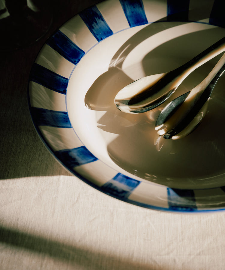The Bestia Serving Set, featuring two metal spoons, rests on a ceramic plate with blue stripes and casts shadows on a light-colored tablecloth in natural light.
