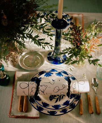 A stylish table features a blue floral plate, cutlery, an abstract white napkin, a blue candle holder, glassware, florals, and Capriccio Holiday Crackers on a light tablecloth—ideal for chic holiday decor.