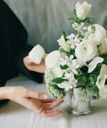 A person’s hand adjusts a small bouquet of white flowers arranged in the Clara Julep Cup on a light-colored surface.