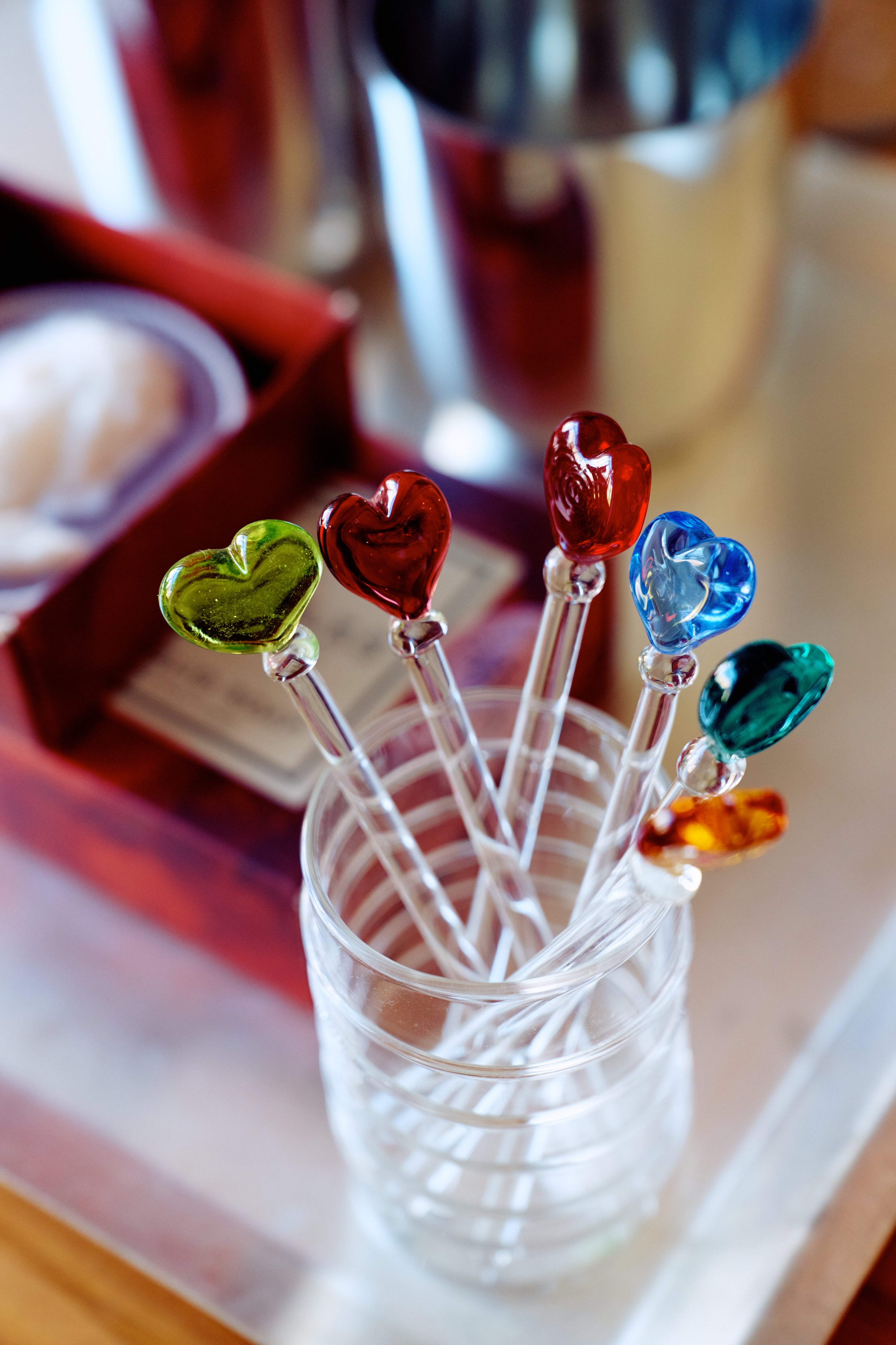 The Heart Cocktail and Nibble Sticks, Set of 6, features clear sticks topped with colorful glass hearts—green, red, blue, and orange—presented in a glass beside a red box and metal cup; perfect accessories for any gathering.
