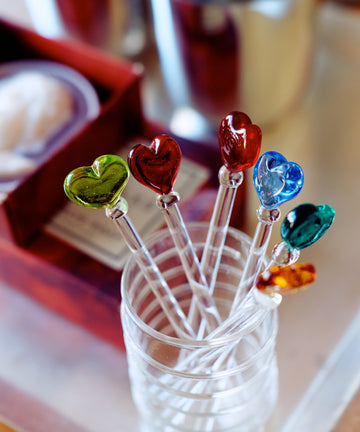 The Heart Cocktail and Nibble Sticks, Set of 6, features clear sticks topped with colorful glass hearts—green, red, blue, and orange—presented in a glass beside a red box and metal cup; perfect accessories for any gathering.