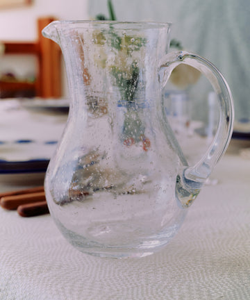 The Lauren Water Jug, a clear French glassware pitcher with condensation, sits on a table set for a meal, with plates and utensils in the background.