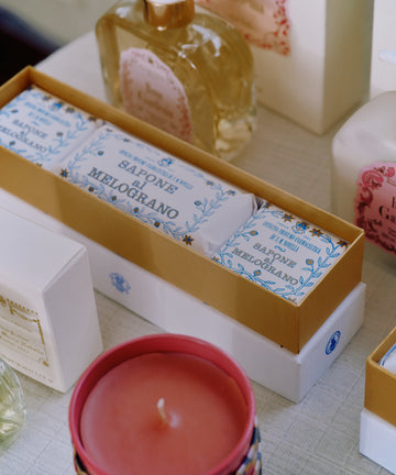 A Melograno Soap Set rests on a table, wrapped in elegant Florentine paper, surrounded by bottles and containers, with a pink candle in the foreground.