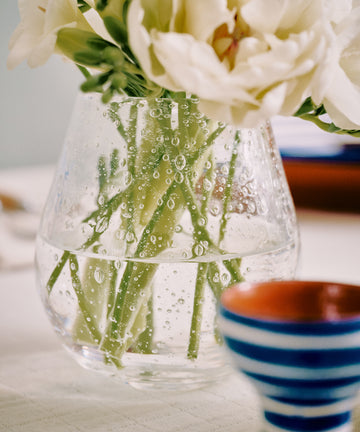 The Lauren Bud Vase, made of clear glass and filled with white flowers and green stems in water with condensation, sits on a table next to a blue and white striped cup.