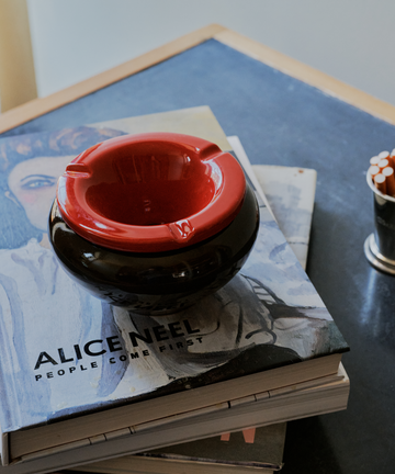 The Mix & Match Ashtray, a chunky ceramic piece handmade in Italy, sits on a dark tabletop atop a stack of books, the top titled Alice Neel: People Come First.