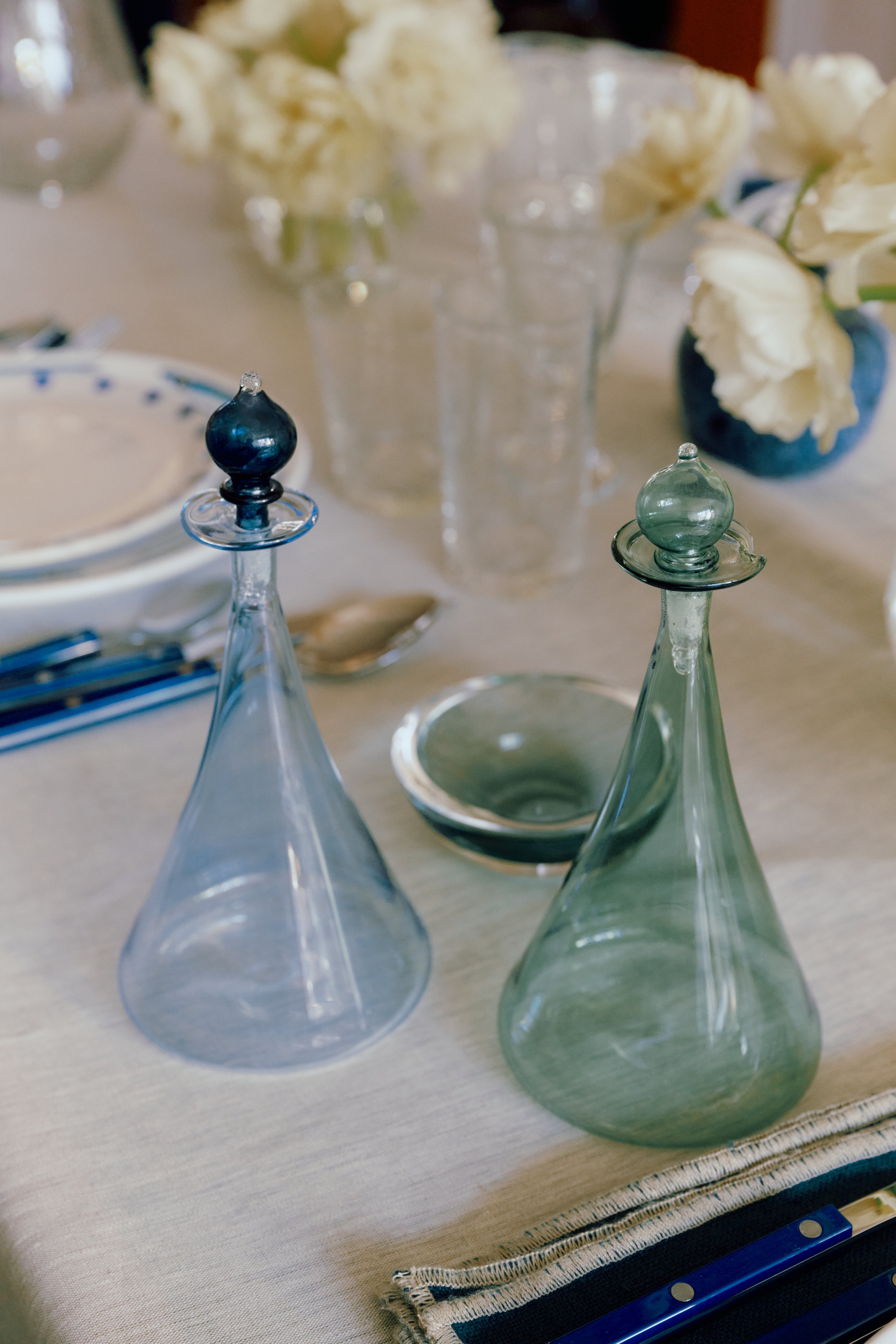 The Duo Vinegar & Oil Flask, a set of clear hand-blown glass decanters with stoppers, sits on a set dining table with blue cutlery, plates, glasses, and white flowers in the background.