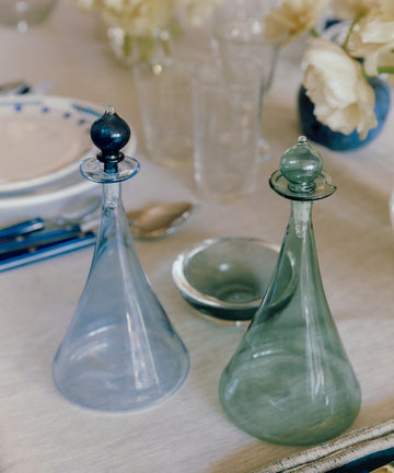 The Duo Vinegar & Oil Flask, a set of clear hand-blown glass decanters with stoppers, sits on a set dining table with blue cutlery, plates, glasses, and white flowers in the background.