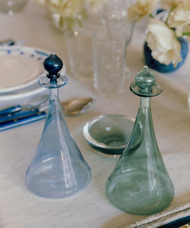 The Duo Vinegar & Oil Flask, a set of clear hand-blown glass decanters with stoppers, sits on a set dining table with blue cutlery, plates, glasses, and white flowers in the background.