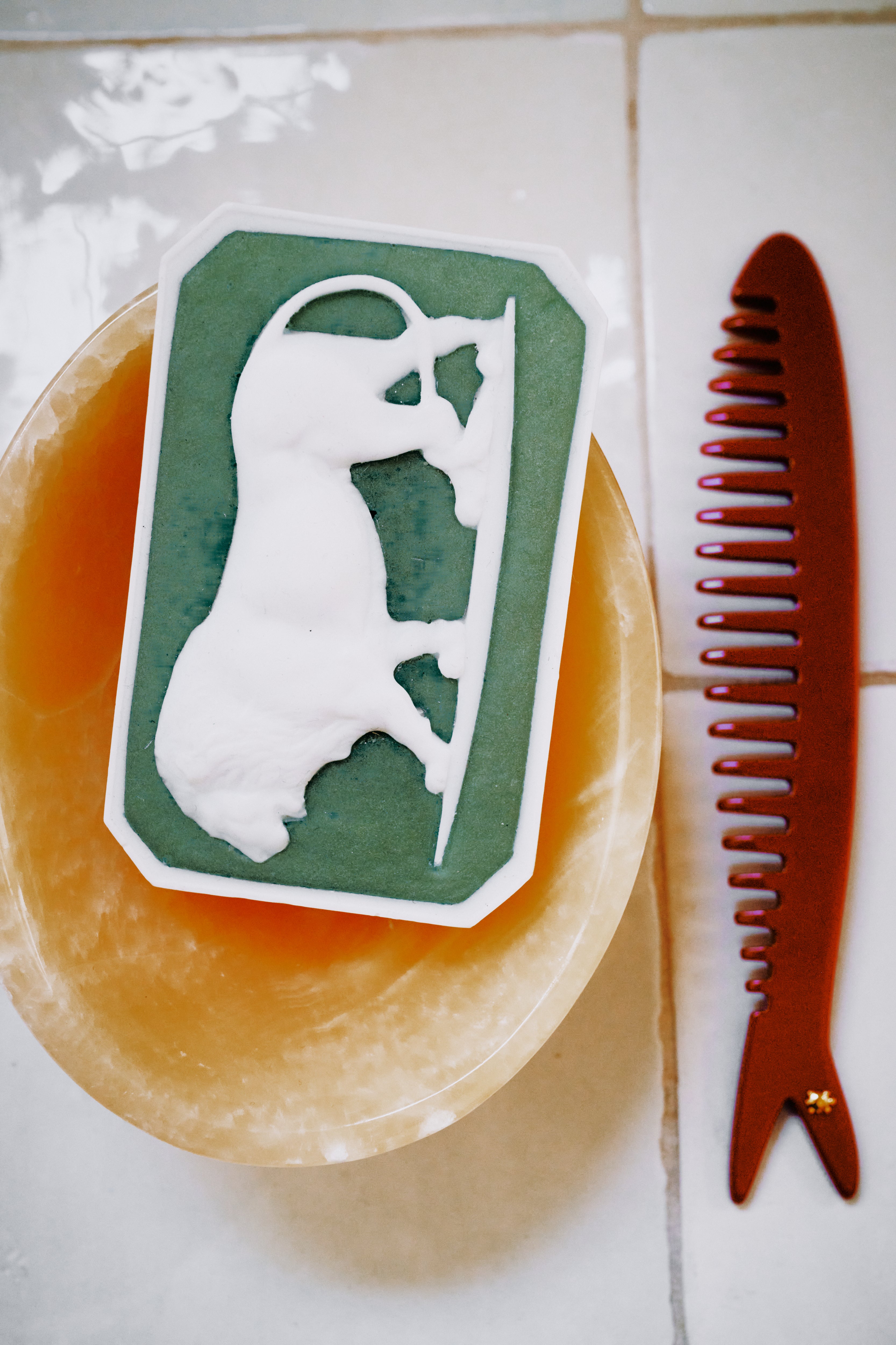 The Clean & Comb Bundle features a green and white rectangular Ancient Lion soap with a raised bull design on a round dish, next to a brown Capri sardine comb, both arranged on a tiled surface.