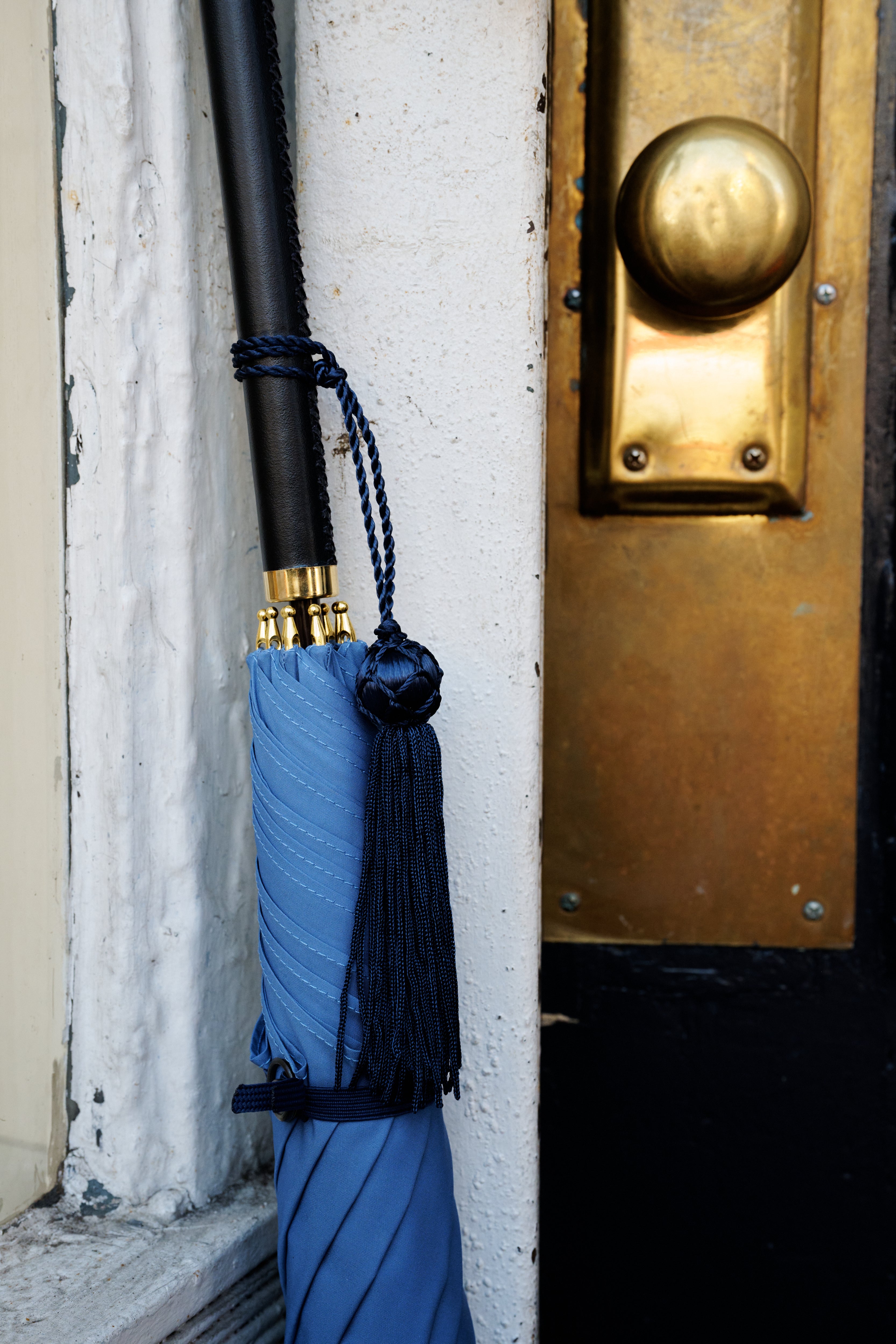 A Victoria Ladies Umbrella with a blue canopy and tassel leans closed against a white wall beside a door featuring a brass handle and lock.