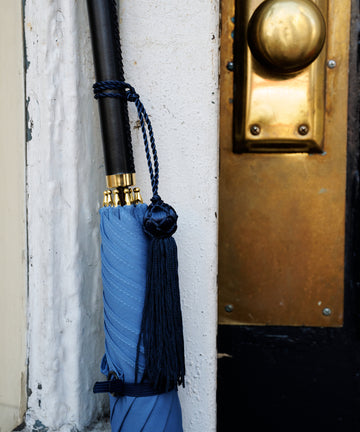 A Victoria Ladies Umbrella with a blue canopy and tassel leans closed against a white wall beside a door featuring a brass handle and lock.