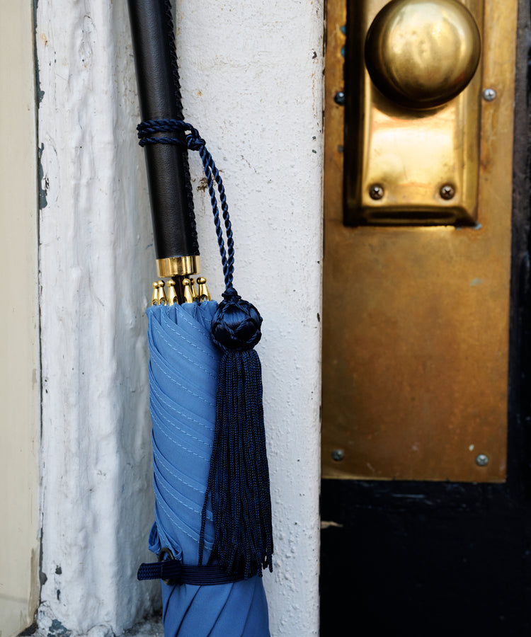 A Victoria Ladies Umbrella with a blue canopy and tassel leans closed against a white wall beside a door featuring a brass handle and lock.
