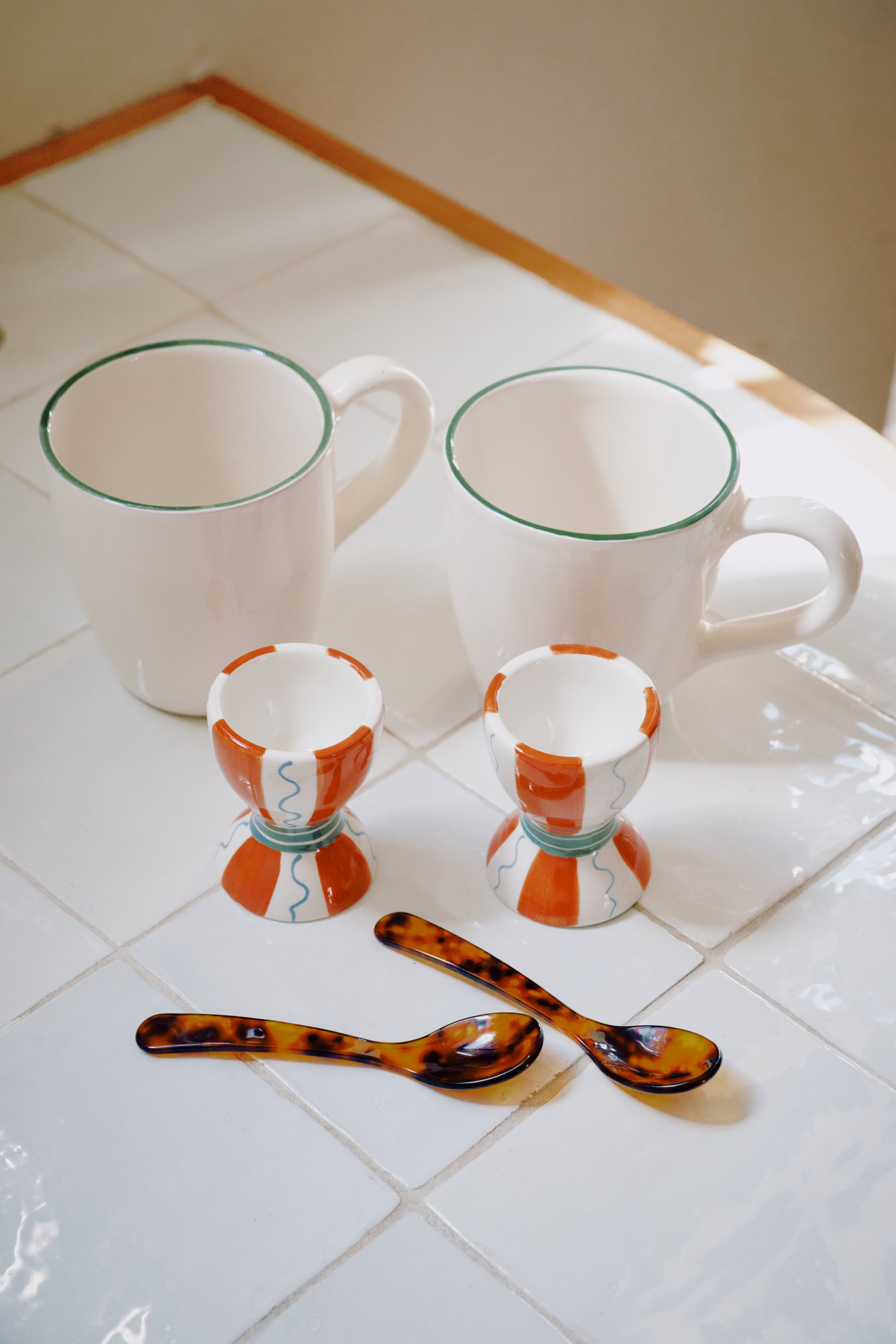 The Breakfast for Two Bundle—two white Guido mugs with green rims, two Wiggle egg cups (orange and blue patterns), and two tortoiseshell Piccolo spoons—rests on a white tiled surface.