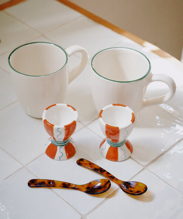 The Breakfast for Two Bundle—two white Guido mugs with green rims, two Wiggle egg cups (orange and blue patterns), and two tortoiseshell Piccolo spoons—rests on a white tiled surface.