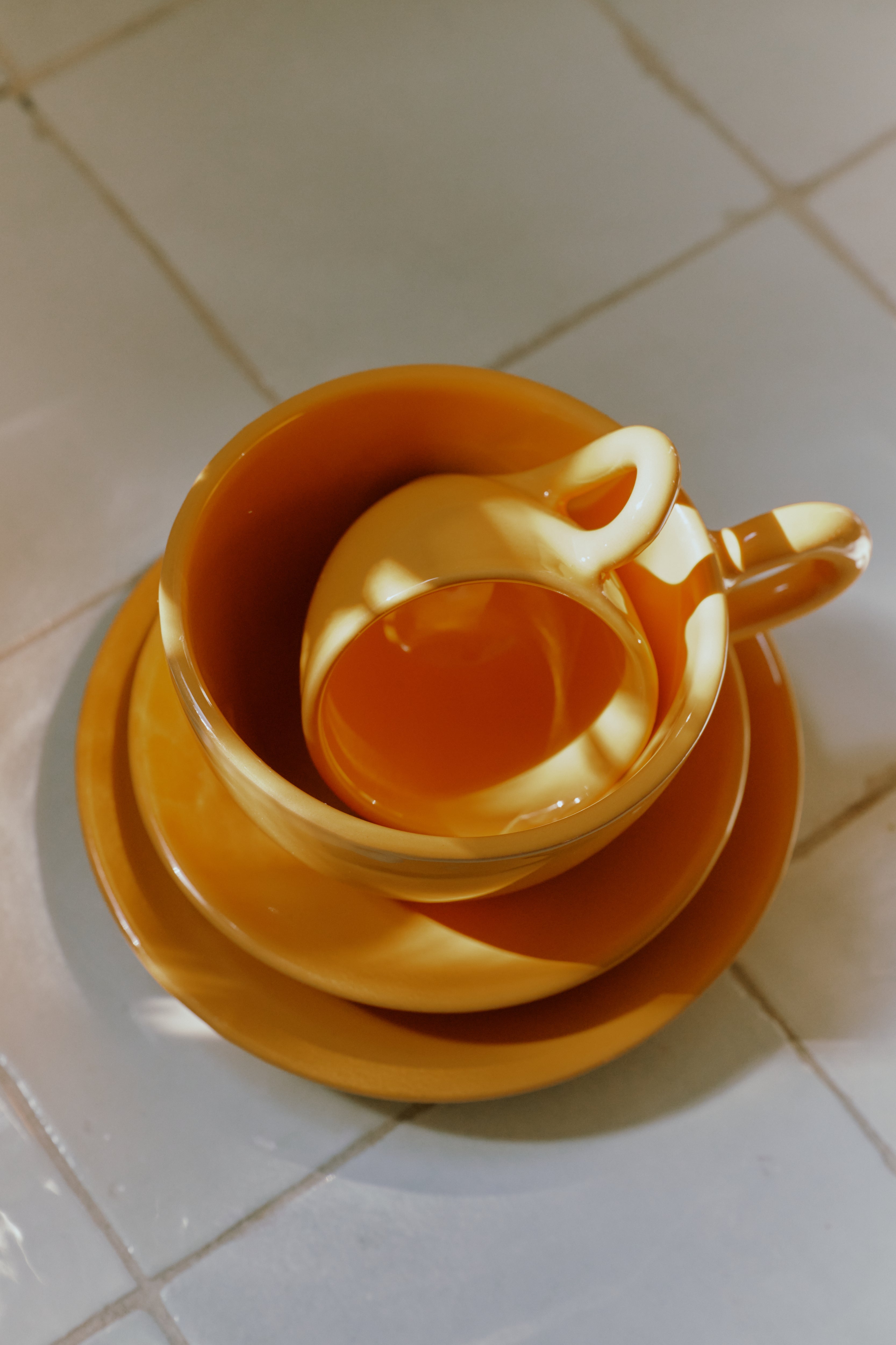 The Gio Espresso Bundle features three stacked yellow ceramic coffee cups and saucers on a tiled surface, with sunlight creating bright reflections and shadows.