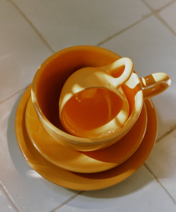 The Gio Espresso Bundle features three stacked yellow ceramic coffee cups and saucers on a tiled surface, with sunlight creating bright reflections and shadows.