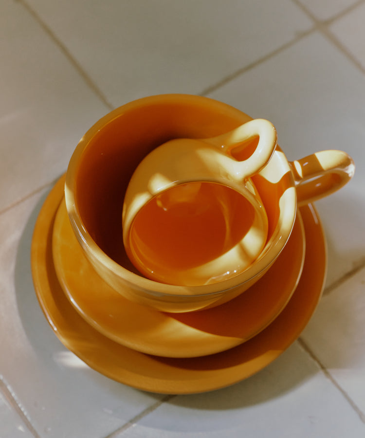 The Gio Espresso Bundle features three stacked yellow ceramic coffee cups and saucers on a tiled surface, with sunlight creating bright reflections and shadows.