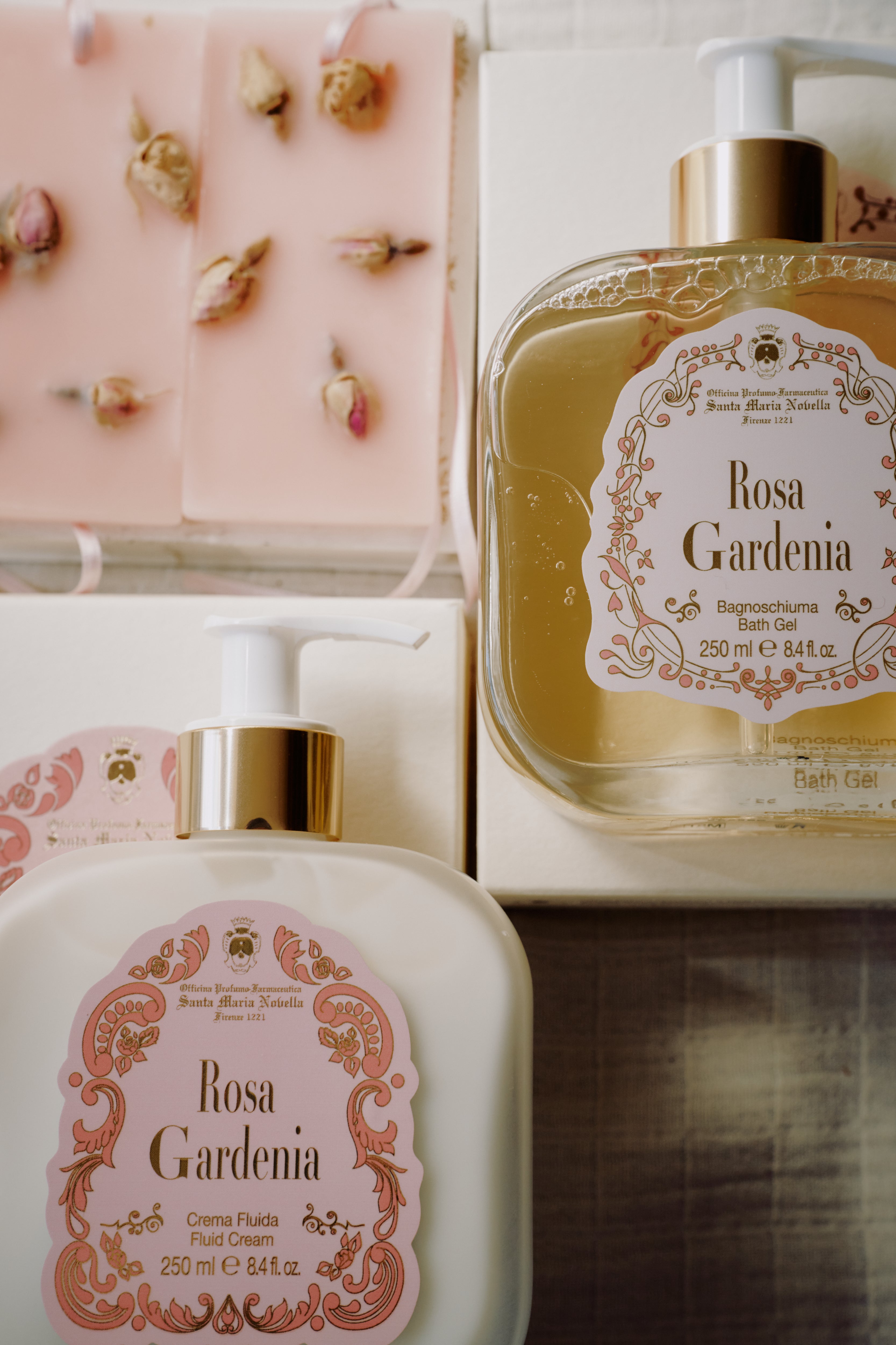 The Rosa Gardenia Bundle features bath gel, fluid cream with decorative labels, and a rosebud-topped pink soap, all filling the air with Rosa Gardenia and sandalwood scents.