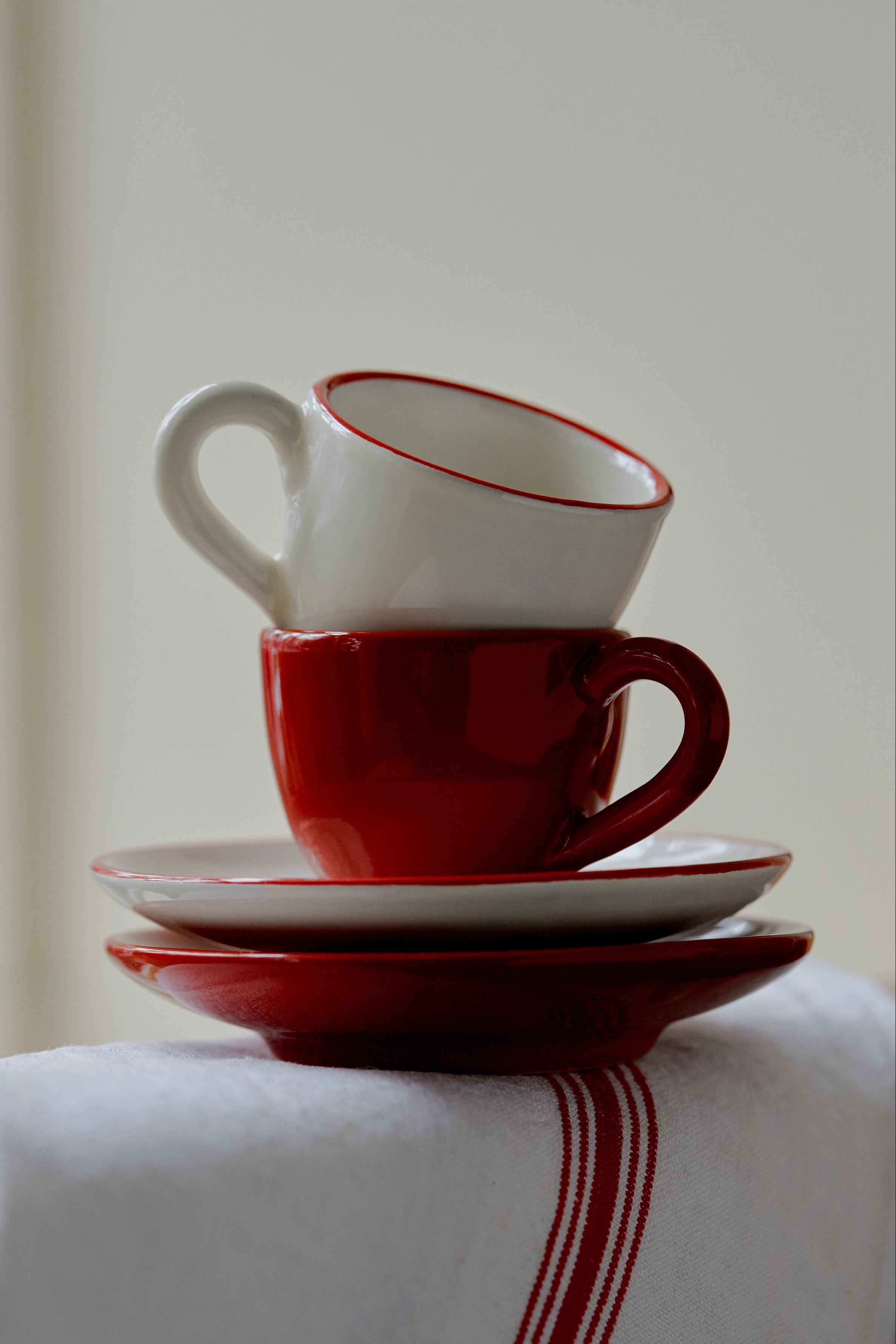 The Gio Espresso Set features two stacked ceramic cups with matching saucers, one in white and one in red, displayed on a white cloth accented with a red stripe.