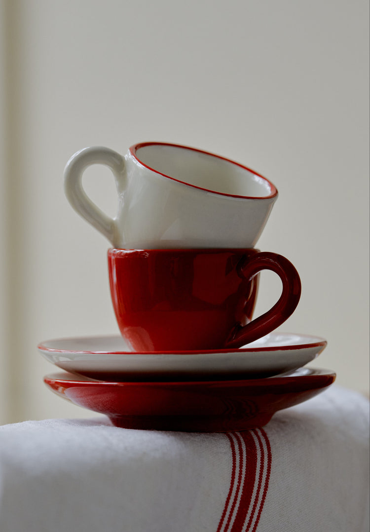 The Gio Espresso Set features two stacked ceramic cups with matching saucers, one in white and one in red, displayed on a white cloth accented with a red stripe.