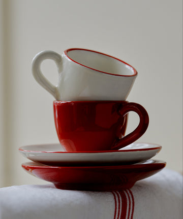 The Gio Espresso Set features two stacked ceramic cups with matching saucers, one in white and one in red, displayed on a white cloth accented with a red stripe.