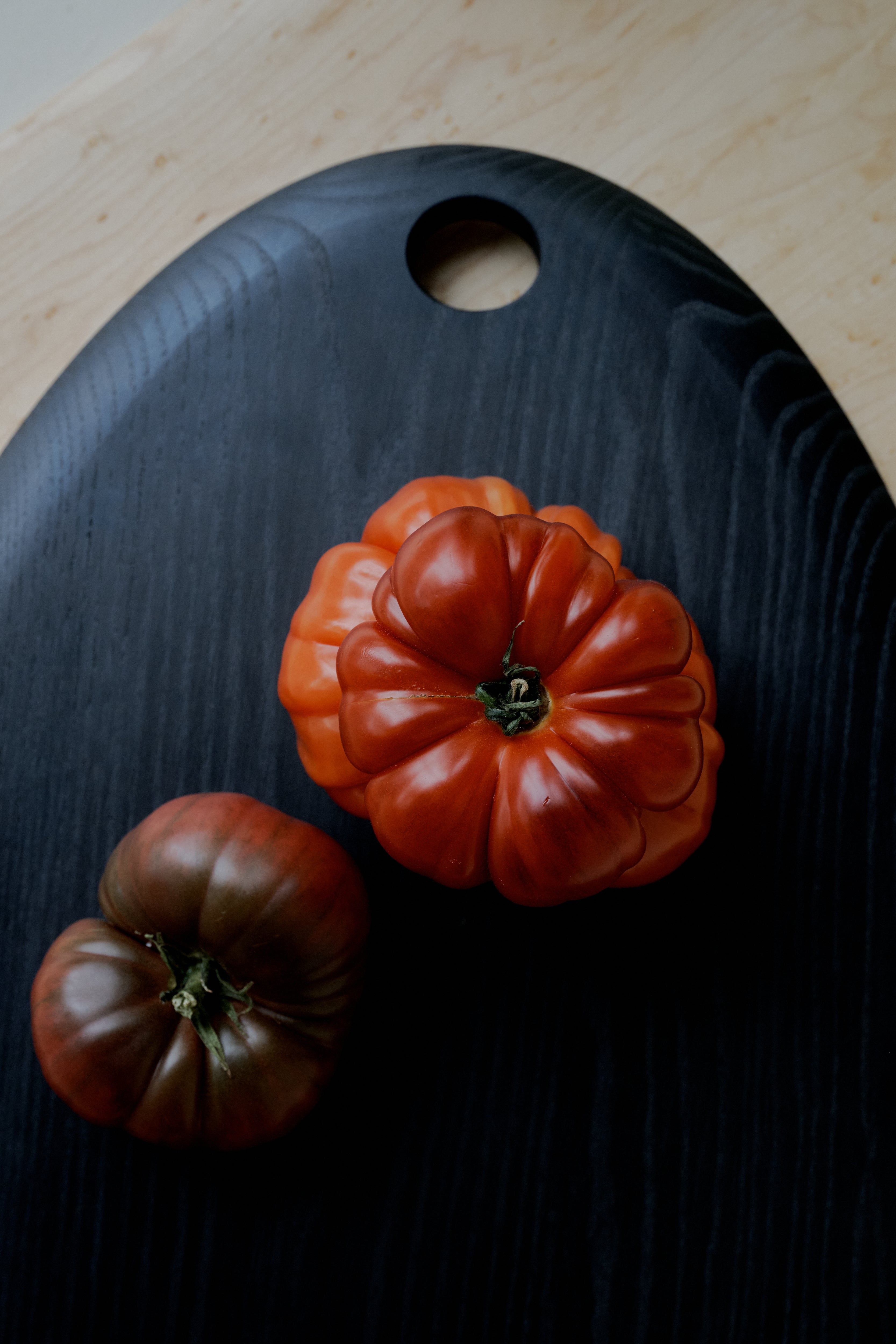Two heirloom tomatoes, one red and one dark reddish-brown, rest on the sleek Alden Egg Board—crafted from black ash and featuring a signature circular hole near the top—creating an elegant centerpiece for any cheese board arrangement.