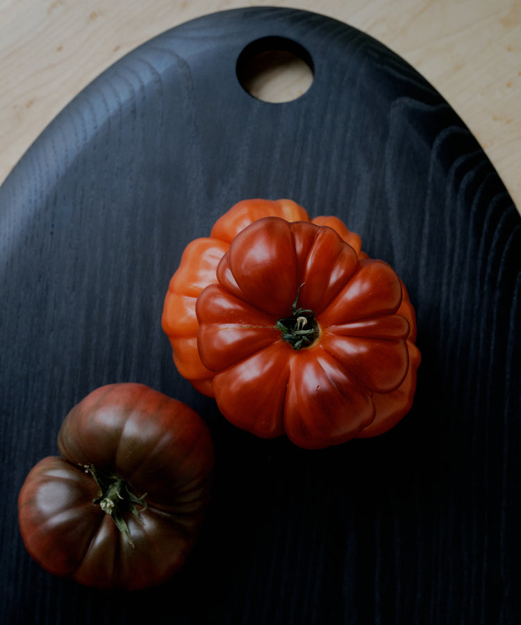 Two heirloom tomatoes, one red and one dark reddish-brown, rest on the sleek Alden Egg Board—crafted from black ash and featuring a signature circular hole near the top—creating an elegant centerpiece for any cheese board arrangement.