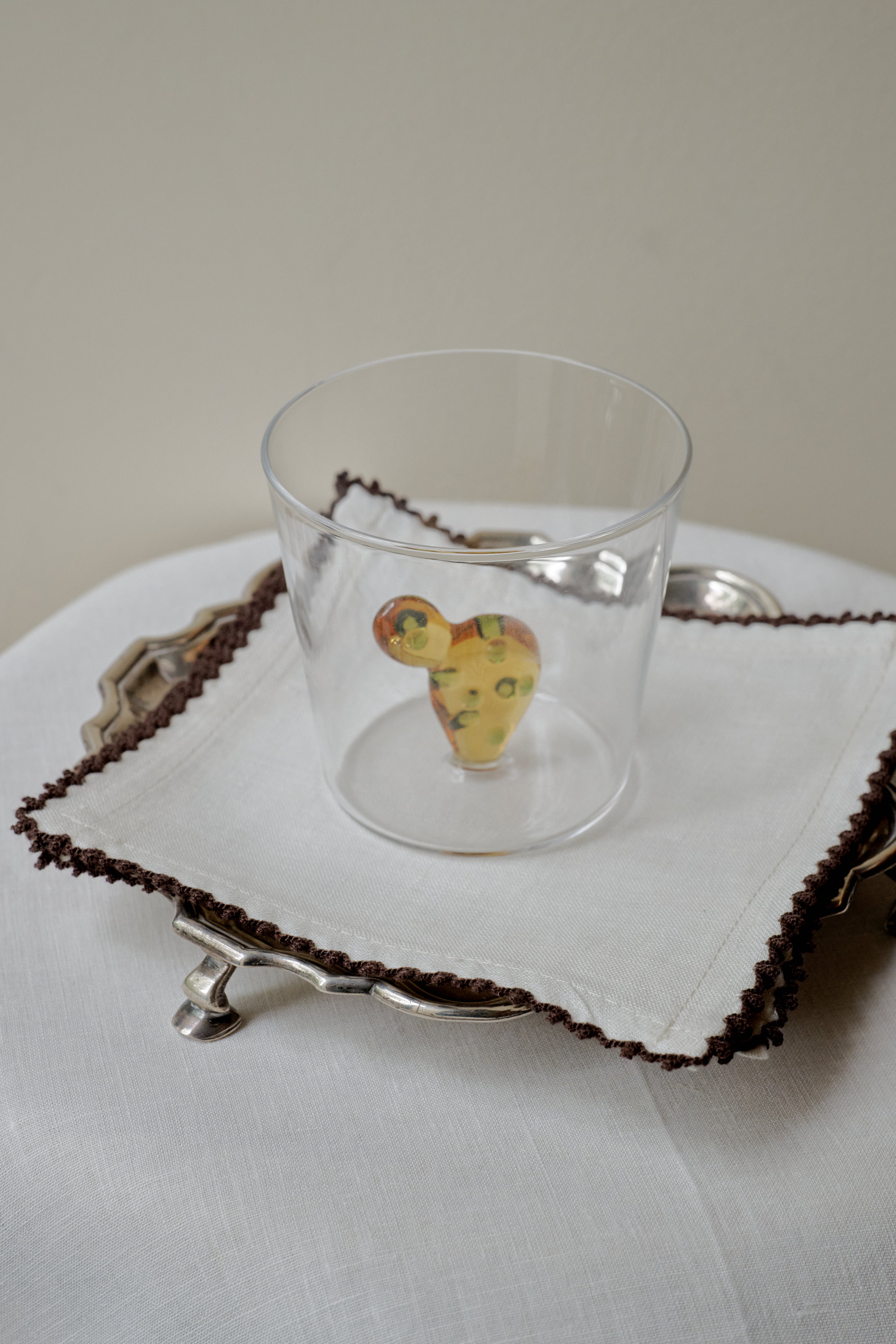 An Agave Tumbler from the set of 4 holds a small, multicolored abstract object. The glass sits on a white napkin with brown trim, neatly arranged on a silver tray.